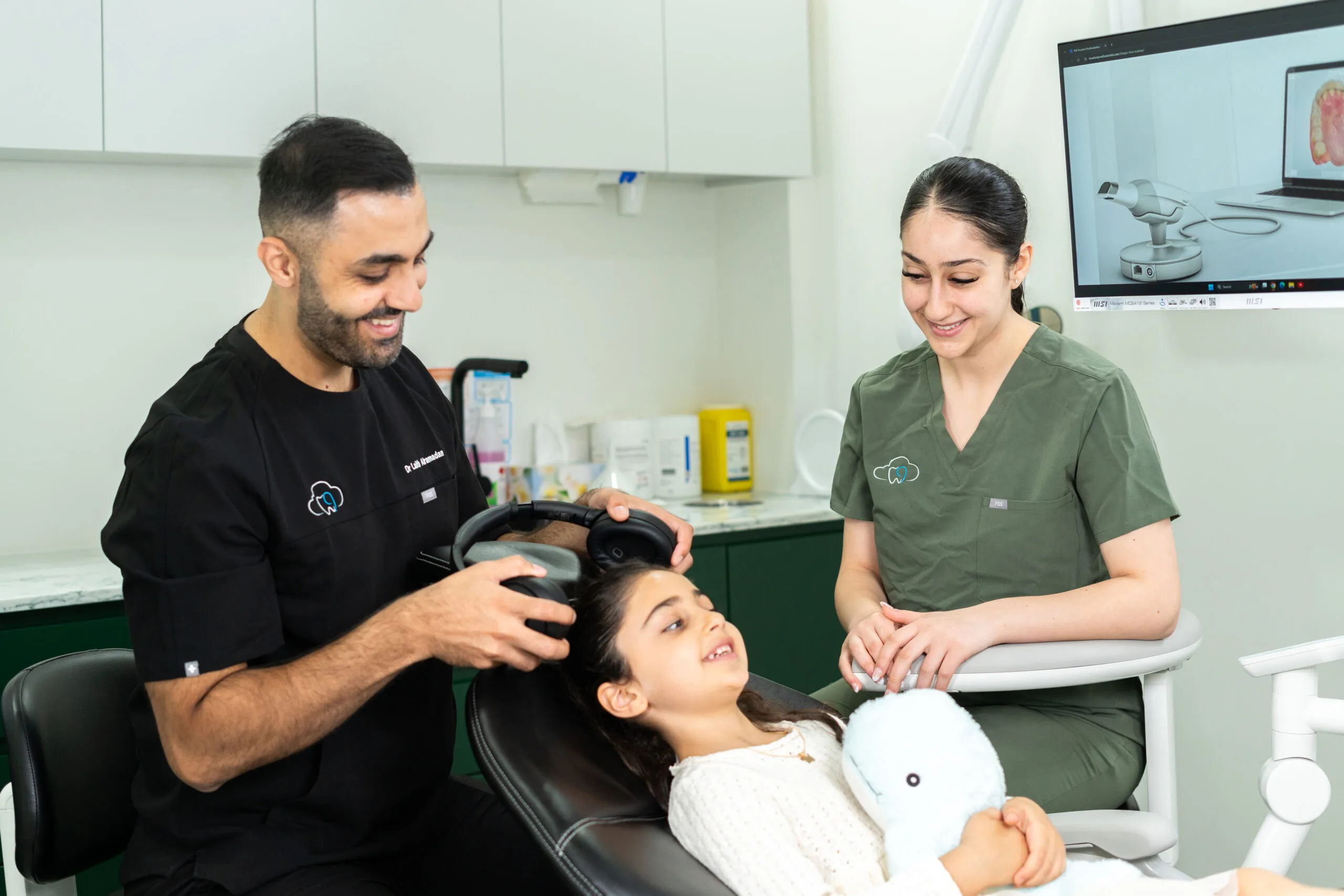 Dentist and dental assistant smiling while helping a young patient relax with headphones during a dental appointment, with a dental imaging screen visible in the background.
